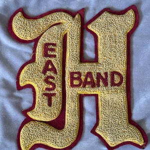 May include: A maroon felt patch with a gold embroidered letter H and the word "BAND" in maroon lettering. The word "EAST" is vertically embroidered in gold on the left side of the patch.