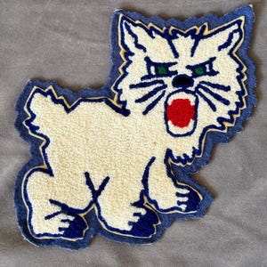 May include: A white felt cat patch with blue and yellow outlining. The cat has a red open mouth and green eyes.