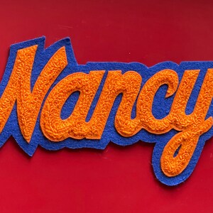 May include: A blue felt patch with the name "Nancy" embroidered in orange yarn.