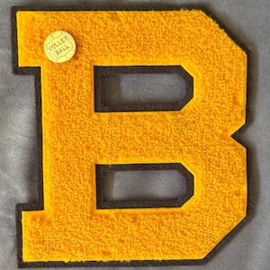 May include: A large, yellow felt letter "B" with a black border and a small, round "Volley Ball" pin. The letter has a textured, fuzzy appearance. The background is a solid, light gray color. This item is likely a patch or emblem.
