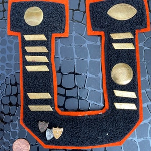 May include: A black felt letter U with an orange border. The letter is decorated with gold-colored athletic pins, including a baseball, football, basketball, and four stripes. There are also two small silver pins with the words "Honor Roll" on them.