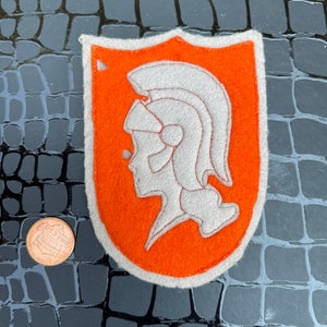 May include: Orange felt patch with a white embroidered image of a Roman soldier's head wearing a helmet.