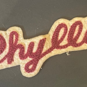 May include: A cream-colored fabric patch featuring the name Phyllis embroidered in a dark red, cursive script. The patch has a die-cut shape that follows the contours of the lettering, with a textured, raised stitch pattern for the name.
