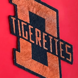 May include: A black and orange felt patch with the word "Tigerettes" in orange lettering.