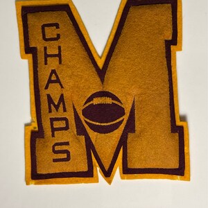 May include: A felt patch with a large gold "M" and a football inside. The "M" has a maroon outline and the football is maroon with a white outline. The word "CHAMPS" is written vertically on the left side of the "M" in maroon.