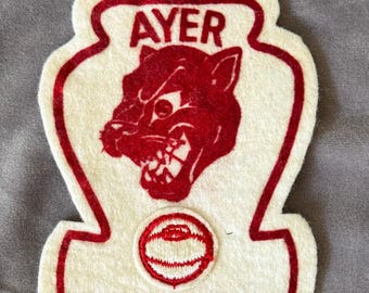 May include: A white felt patch with a red border and a red panther graphic. The word "AYER" is at the top. A red circle with a design is at the bottom. The patch is on a gray surface.