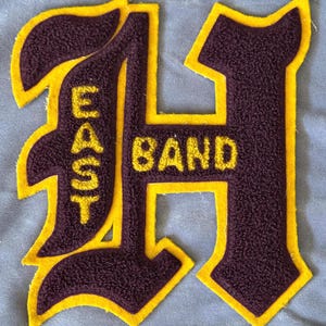 May include: A maroon felt patch with a yellow outline in the shape of the letter H. The word "BAND" is written across the top of the H and the word "EAST" is written vertically down the left side of the H.