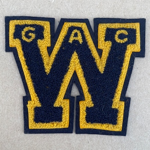 May include: A black felt letter W with a yellow outline. The letters G, A, and C are sewn into the top of each side of the letter W.