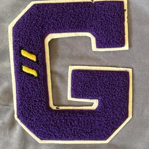 May include: Purple felt letter G with a white outline and two yellow stripes. The letter is sewn onto a light purple fabric.