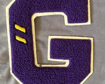 May include: Purple felt letter G with a white outline and two yellow stripes. The letter is sewn onto a light purple fabric.