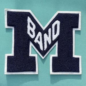 May include: A navy blue letter "M" patch with the word "BAND" in white, stitched onto a light teal fabric. The patch has a white border. The image is a close-up of the patch.