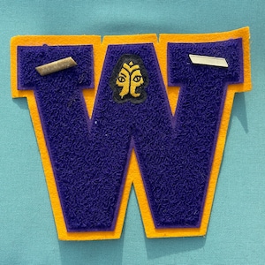 May include: A purple felt letter W with a yellow border. The letter is decorated with a gold and black emblem with two faces.