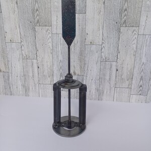 May include: A metal sculpture with a long, thin, vertical piece and a base with three circular pieces. The sculpture is made of metal and has a dark gray color.