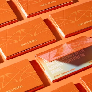 Puede incluir: Primer plano de varias cajas naranjas con letras doradas que dicen "SMKN GODDESS". Las cajas están dispuestas en un patrón escalonado sobre una superficie naranja claro. Una caja está abierta, revelando una tarjeta blanca y amarilla con el texto "ARE DESIGNED WITH SOPHISTICATION IN MIND. ALLURING FLAVORS AND AROMAS @SMKNGODDESS".