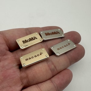 Könnte beinhalten: Vier rechteckige Metallanhänger mit abgerundeten Ecken. Zwei sind goldfarben und zwei silberfarben. Auf den goldenen Anhängern steht "MoMA" und "macac", auf den silbernen Anhängern "MoMA" und "macac". Jeder Anhänger hat kleine Nieten an jeder Ecke.