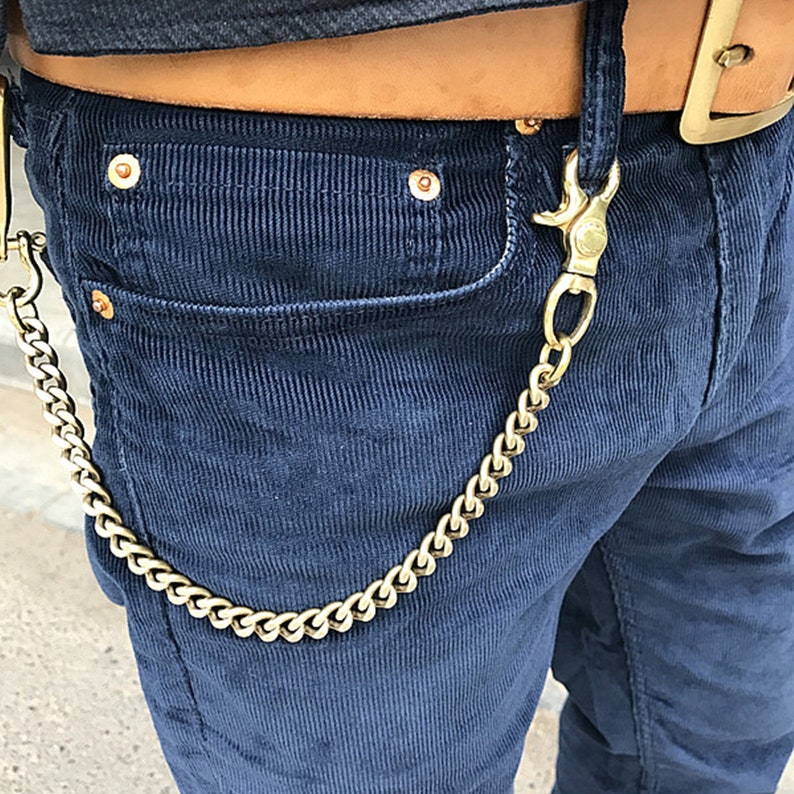 May include: A gold-colored wallet chain attached to a pair of navy blue corduroy pants. The chain is connected to a belt loop and a pocket. The belt is brown leather with a gold buckle.