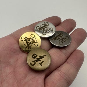 May include: Five metal buttons in gold, silver, and bronze. Each button has a different design, including a skull and crossbones and a lightning bolt with letters. The buttons are round with a slightly raised edge. The buttons are held in a hand.