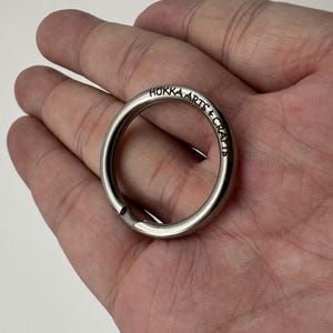 Puede incluir: Un llavero plateado con el texto "Hukka Arts + Crafts" grabado en la superficie exterior. El llavero es circular con una pequeña abertura para sujetar las llaves. El llavero se sostiene en una mano abierta.