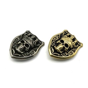 Könnte beinhalten: Zwei metallene Schild-förmige Knöpfe mit Totenkopf und Krone. Ein Knopf ist silberfarben und der andere goldfarben.