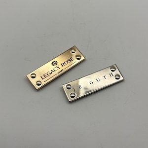 Peut inclure: Deux étiquettes rectangulaires en métal. L'une est dorée avec le texte "LEGACY ROSE TEXAS" et un emblème de rose. L'autre est argentée avec le texte "JE GUTH". Les deux étiquettes ont quatre rivets.