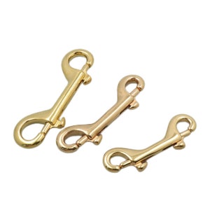 May include: Three gold-colored bolt snap hooks of varying sizes are arranged on a white background. Each hook has a spring-loaded clasp and a swivel eye. The hooks are made of a shiny, metallic material.