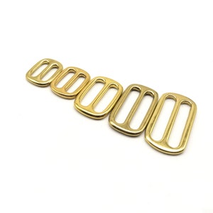 Può includere: Un set di sei fibbie scorrevoli in metallo dorato di diverse dimensioni. Le fibbie sono rettangolari con due fori ovali al centro.