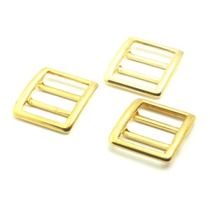 May include: Three gold-colored rectangular slider buckles with rounded corners. Each buckle has three parallel bars across the center. The buckles are made of metal and are displayed against a white background.