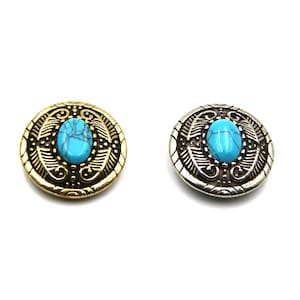 Op de afbeelding: Twee ovale metalen knopen met een turquoise steen in het midden. De knopen hebben een sierlijk ontwerp rond de steen. Eén knop is goud en de andere is zilver.