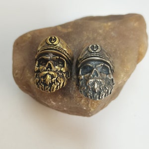 Op de afbeelding: Twee schedelvormige bedels met een baard en een pet. De bedel aan de linkerkant is goud en de bedel aan de rechterkant is zilver.