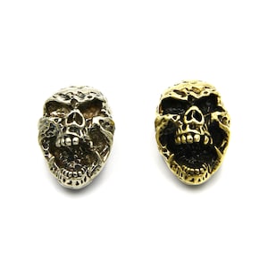 Puede incluir: Dos botones de metal con forma de cráneo, uno plateado y otro dorado, con tallas detalladas y bocas abiertas.