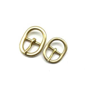 Puede incluir: Dos hebillas de latón de forma ovalada, una más grande que la otra. Ambas hebillas tienen un acabado liso y pulido y un diseño simple y clásico.