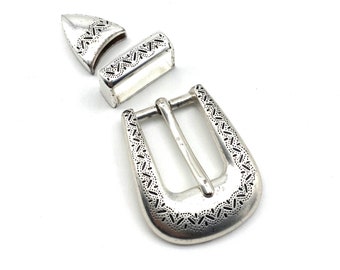 May include: Silver belt buckle with a decorative pattern. The buckle is in three pieces, a main buckle, a top piece, and a side piece.