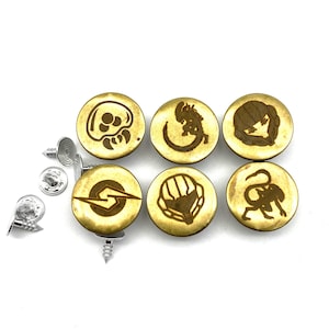 Puede incluir: Seis botones de metal de color dorado con diferentes diseños grabados. Los diseños incluyen una criatura de la película Alien, una calavera, un personaje de videojuego, un casco y un logo. Los botones están sobre un fondo blanco.