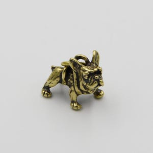 Op de afbeelding: Een kleine, gedetailleerde messing figuur van een Franse Bulldog. De hond wordt afgebeeld in een staande pose met zijn oren omhoog. Het messing heeft een warme, gouden tint en de figuur heeft een ring aan de achterkant, wat suggereert dat het een bedel of hanger kan zijn.