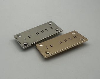 Puede incluir: Dos etiquetas rectangulares de metal, una plateada y otra dorada, cada una con el texto "JE GUTH" en negro. Las etiquetas están aseguradas con cuatro pequeños remaches en cada esquina. La etiqueta plateada está encima de la dorada.