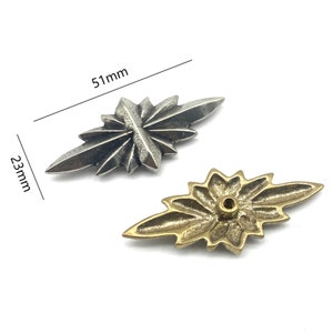 Op de afbeelding: Twee metalen stervormige decoratieve versieringen. Eén is zilver en meet 5,1 cm bij 2,3 cm. De andere is goud en meet 5,1 cm bij 2,3 cm.