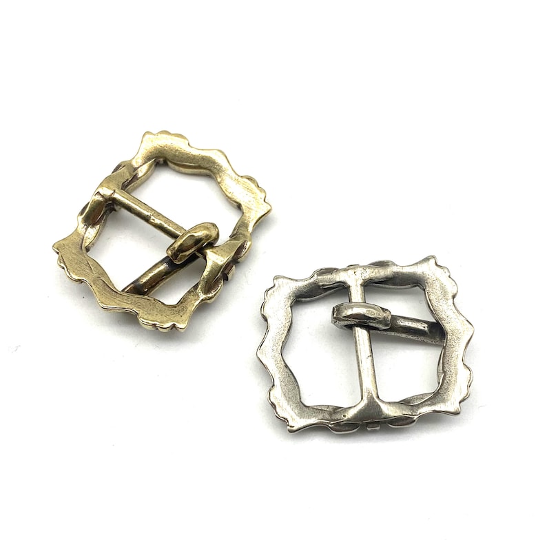 May include: Two ornate metal belt buckles, one gold and one silver, with a square shape and intricate detailing around the edges.