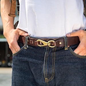 Peut inclure: Une ceinture en cuir marron avec une boucle en laiton et une fermeture à crochet. La ceinture est portée sur un jean bleu.