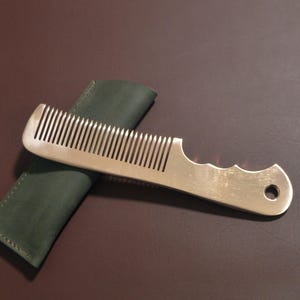 Può includere: Un pettine di metallo con un manico e un foro all'estremità, appoggiato su un astuccio in pelle verde scuro. Il pettine ha denti fitti e un manico curvo per l'impugnatura. Lo sfondo è una superficie marrone scuro.