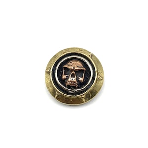 Op de afbeelding: Een gouden knop met een zwarte en zilveren ring rond een koperen schedelontwerp.