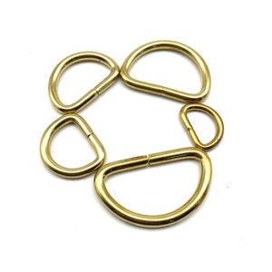 May include: Five gold-toned metal D-rings of varying sizes. The rings are arranged in a circle.