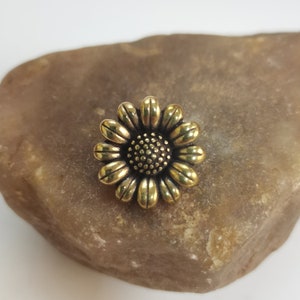 Könnte beinhalten: Eine detaillierte Sonnenblume aus goldfarbenem Metall. Die Blume hat mehrere Blütenblätter, die ein strukturiertes Zentrum umgeben. Die Sonnenblume ist ein dekoratives Element, möglicherweise für Schmuck oder Bastelarbeiten.