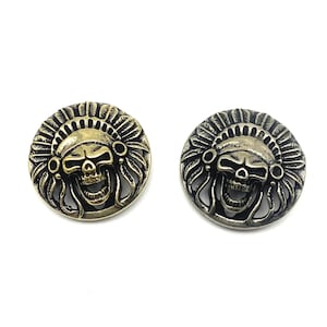 Puede incluir: Dos botones de metal de color dorado con un cráneo que lleva un tocado. Los botones son redondos y tienen un diseño en relieve.
