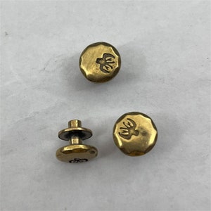 May include: Three gold-toned metal buttons with a raised design. Two buttons are round and one button is a two-piece design with a post and a round top.