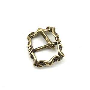 May include: A gold-toned metal buckle with a decorative design. The buckle has a square shape with rounded corners and intricate details.