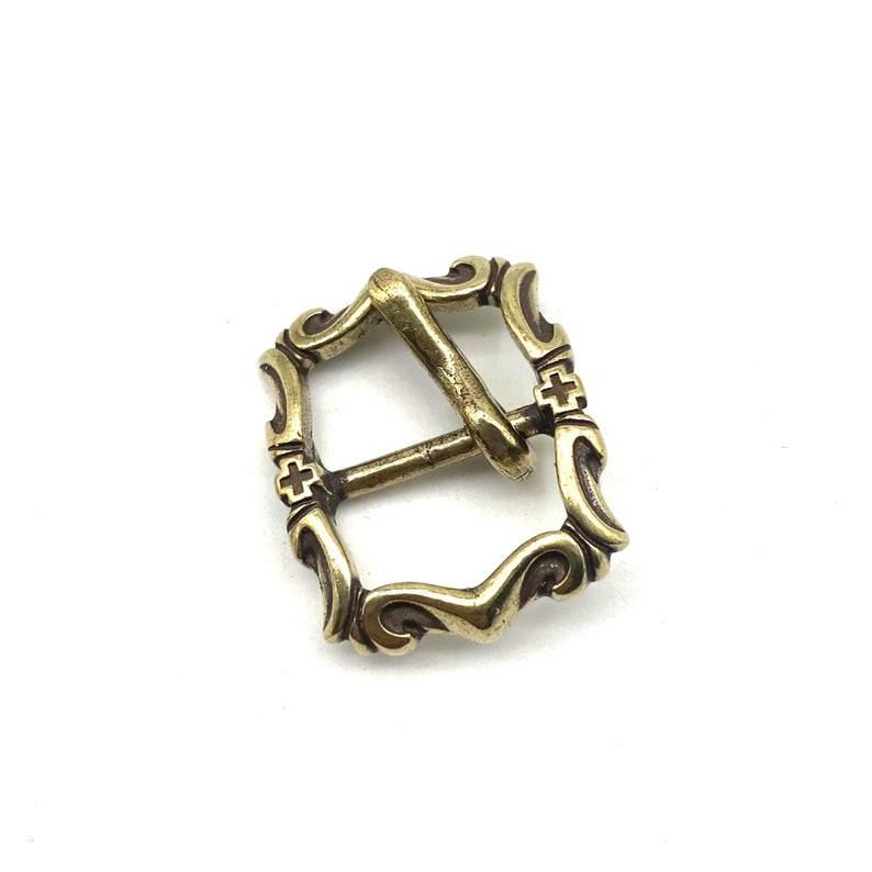 May include: A gold-toned metal buckle with a decorative design. The buckle has a square shape with rounded corners and intricate details.