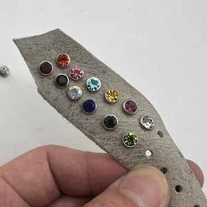 May include: A grey leather strap adorned with a selection of colourful, round rhinestones. The rhinestones are in shades of red, pink, blue, yellow, and clear. A silver stud is visible to the left of the strap. The strap has several holes.