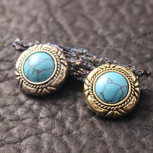 Pode incluir: Duas pedras turquesa cabochon engastadas em metal dourado com detalhes em forma de corda ao redor das bordas.