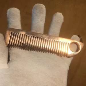 May include: A gold-colored metal comb with a round hole at the end. The comb is lying on a white surface.