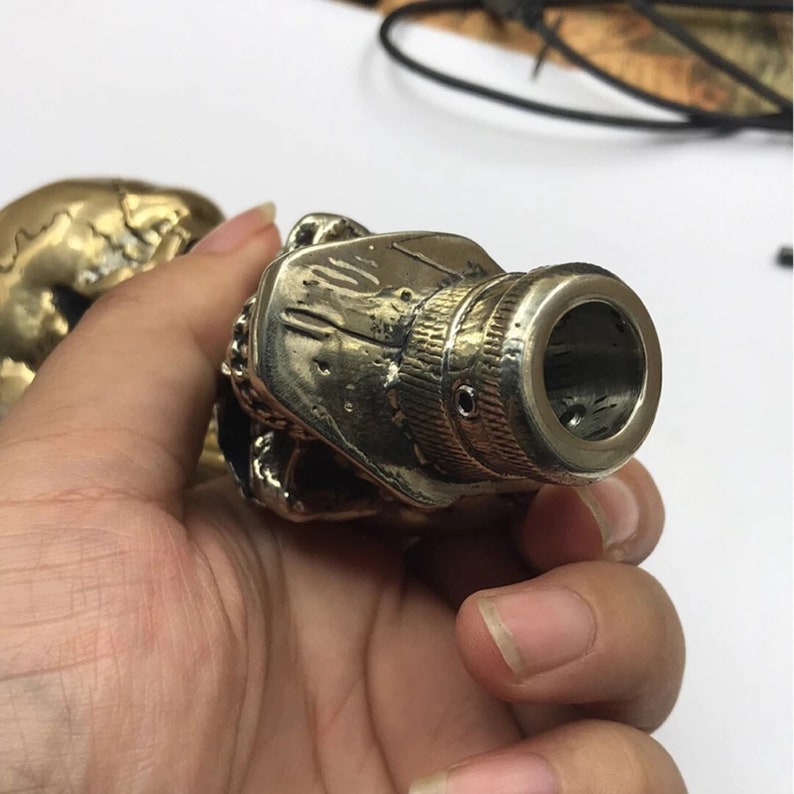 May include: A brass skull-shaped gear shift knob with a detailed design. The knob has a threaded end for attaching to a gear shift lever.